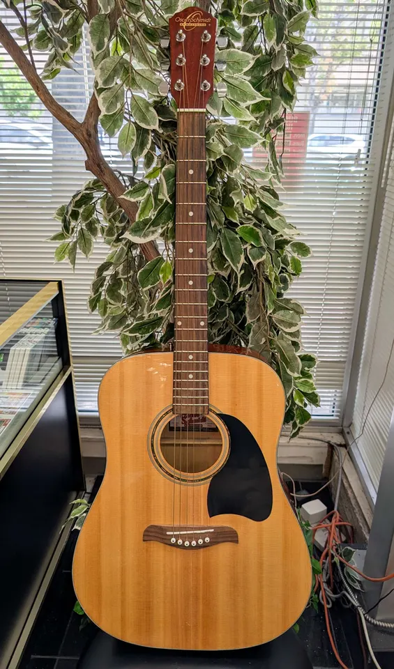Natural Oscar Schmidt by Washburn OG2TPAK dreadnought acoustic guitar showing its spruce top, black pickguard, rosewood bridg