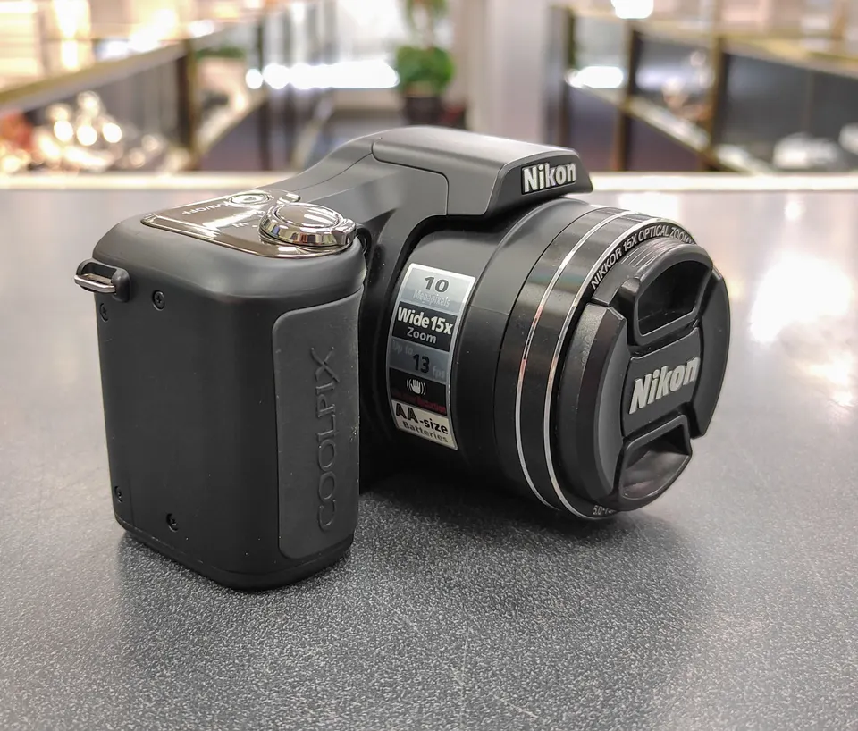 Nikon COOLPIX L100 black digital camera shown from multiple angles with lens cap, pop-up flash, LCD screen, battery sd card